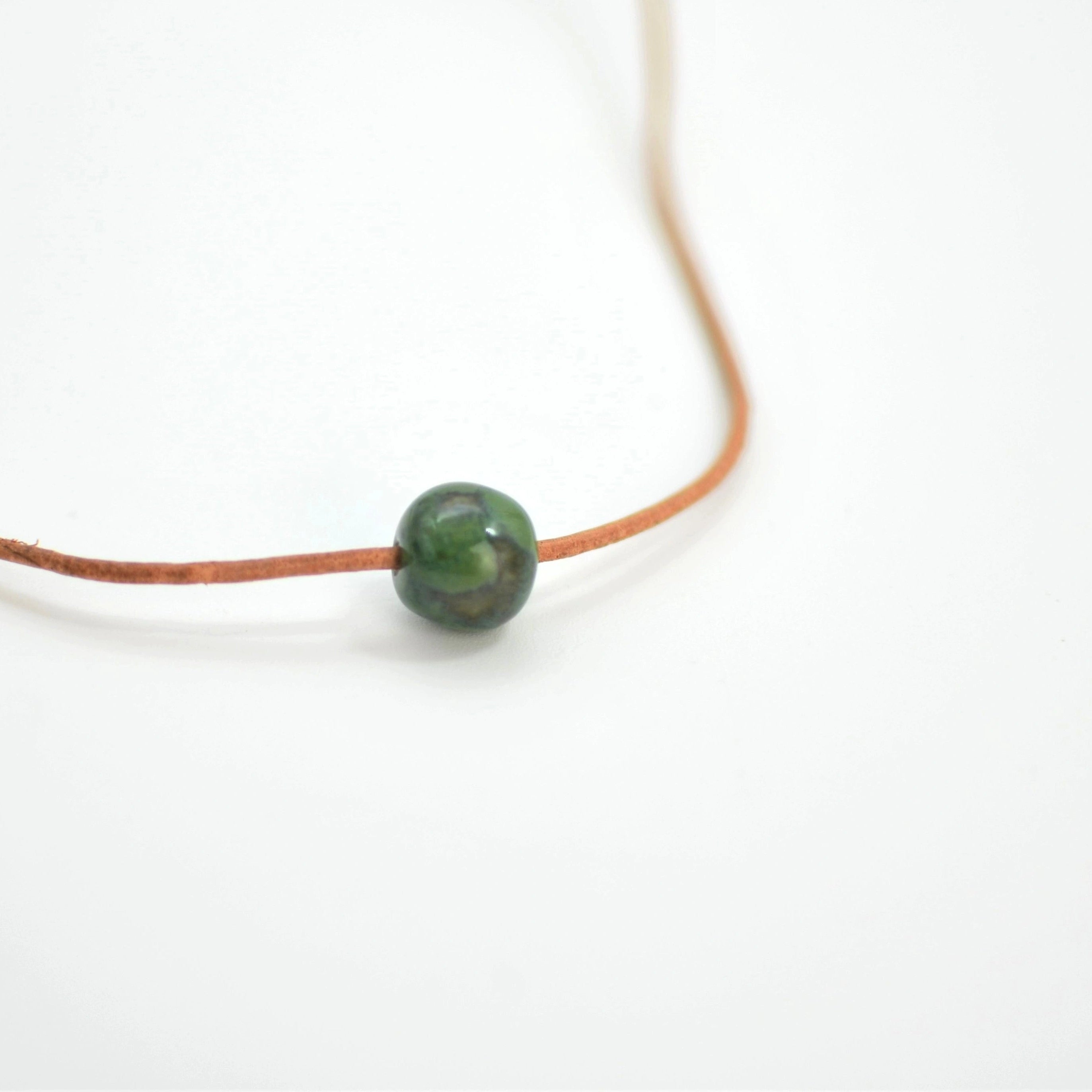 Collar de cerámica esfera verde azulado sobre superficie natural, joyería artesanal única de Maia Cerámica