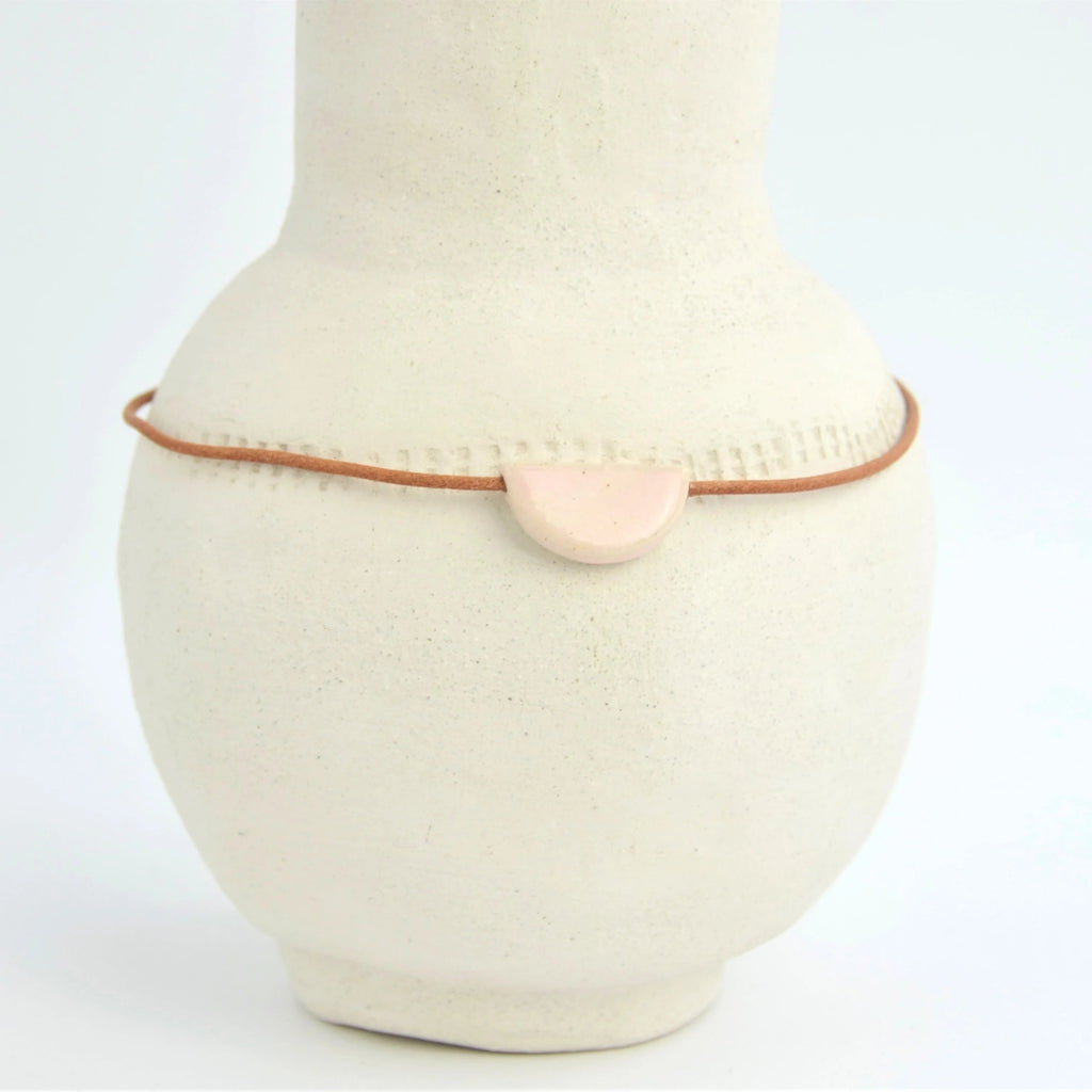 Collar de cerámica media luna rosa sobre superficie natural, joyería artesanal delicada hecha a mano por Maia Cerámica

